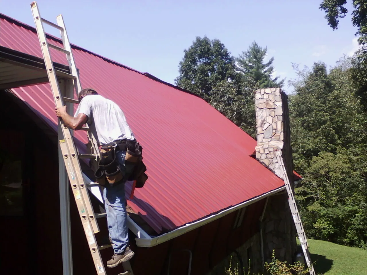 Roofer on a ladder beside a red metal roof and stone chimney — Soffit & Fascia Repair by Roofing Brothers in Chatham